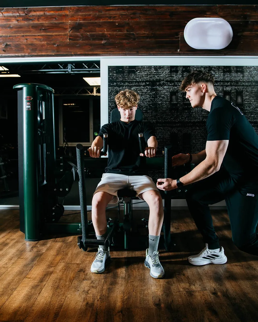 Ein Trainer kniet neben einem jungen Mann und stellt ihn am Kraftgerät ein.
