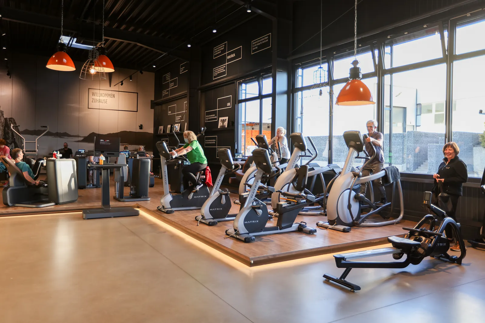 Cardiobereich im effectiv mit Crosstrainern und Rudergerät auf Holzboden, darüber orangefarbene Pendelleuchten und eine große Fensterfront.