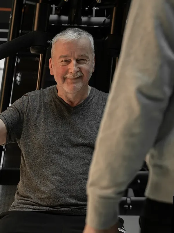 Ein älteres Mitglied lächelt am Kraftgerät, ein Trainer steht daneben.